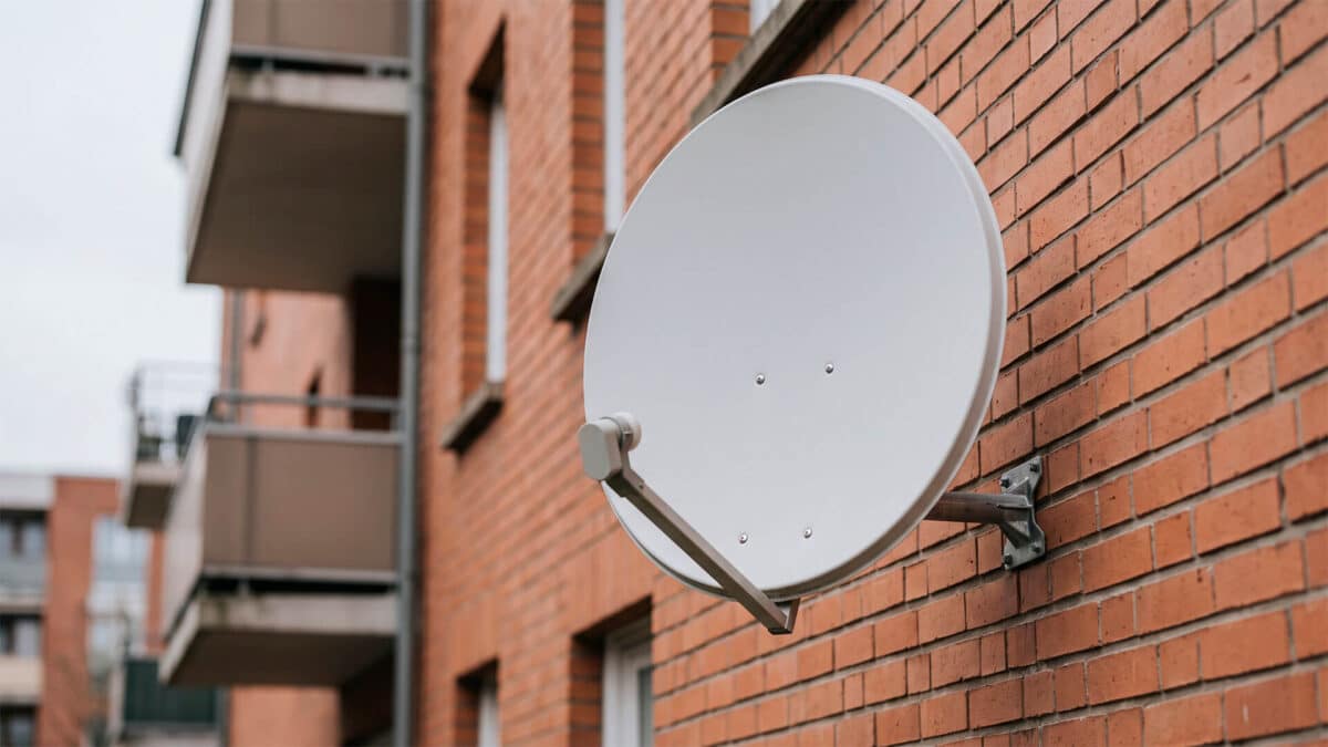 Antena parabólica blanca instalada en la fachada de ladrillo de una comunidad de vecinos.