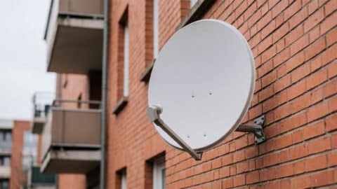 Antena parabólica blanca instalada en la fachada de ladrillo de una comunidad de vecinos.