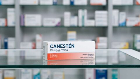 Caja de Canesten crema sobre el mostrador de cristal de una farmacia con estanterias de medicamentos al fondo