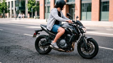 Motorista con casco, sudadera y pantalón corto conduciendo una motocicleta moderna por una calle urbana.
