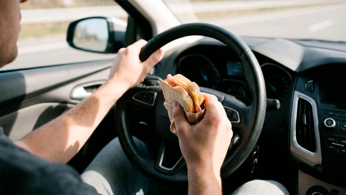 Conductor sujetando un sándwich contra el volante mientras conduce por carretera.
