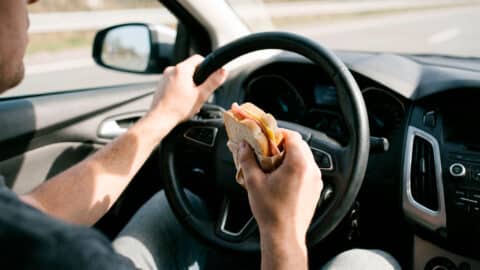 Conductor sujetando un sándwich contra el volante mientras conduce por carretera.