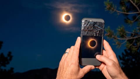 Manos sosteniendo un móvil con filtro solar protector en la cámara para grabar un eclipse solar en el cielo.
