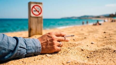 Mano sosteniendo un cigarrillo encendido en la arena frente a una señal de prohibido fumar en la playa.