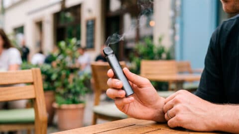 Mano sosteniendo un vaper emitiendo vapor en la terraza de una cafetería al aire libre.