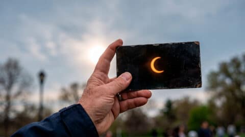 Mano sosteniendo un cristal de soldador sombra 14 a través del cual se observa un eclipse solar parcial en el cielo.